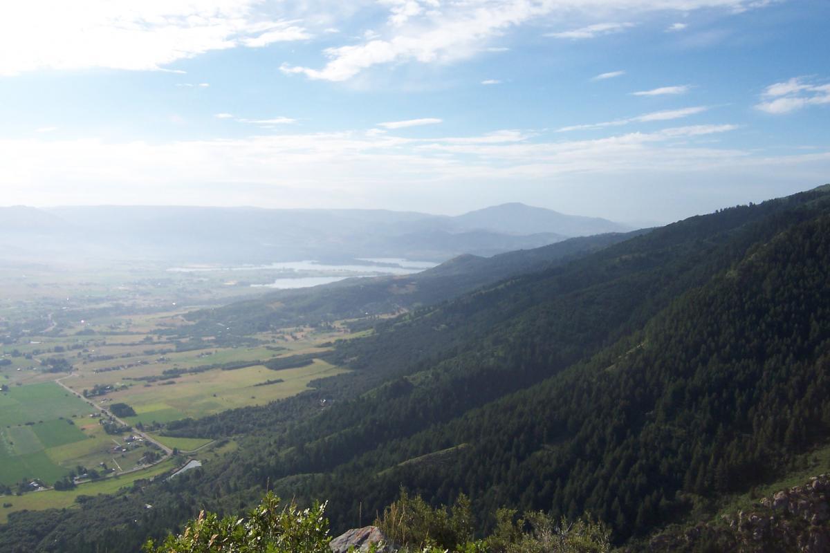 A panoramic view of rolling hills and valleys under a clear blue sky, with patches of green farmland and a winding road visible in the foreground. In the distance, a body of water reflects the sky, surrounded by forested mountains. Northern Skyline mountain bike trail.
