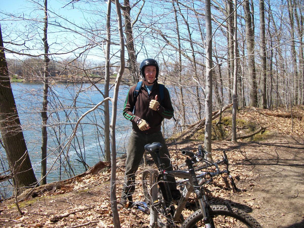 A person wearing a helmet and gloves stands beside a mountain bike along a wooded trail near a river. The background features bare trees and a clear blue sky, indicating early spring. The individual is smiling and giving a thumbs-up, suggesting a sense of enjoyment in outdoor biking. Great Bear mountain bike trail.