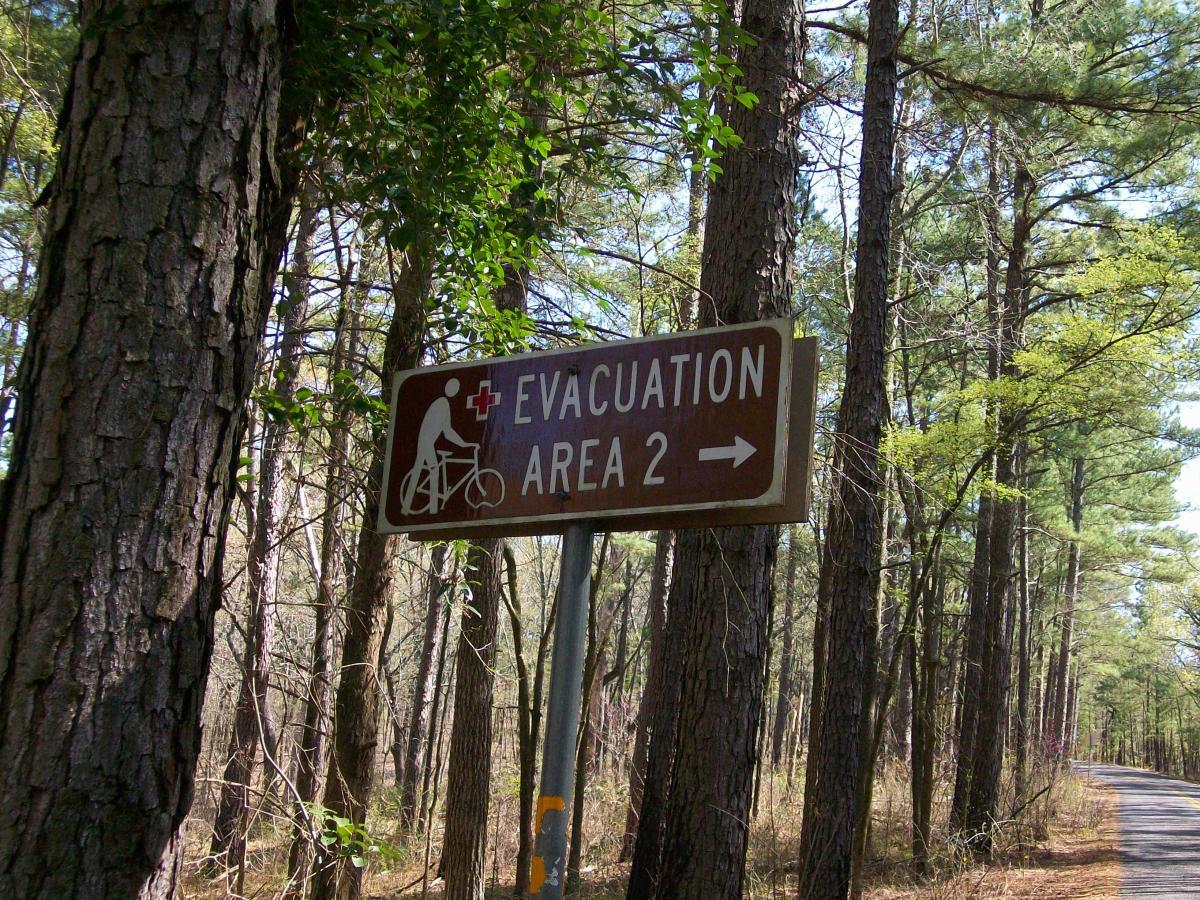 Signpost indicating "Evacuation Area 2" with a graphic of a person riding a bicycle, surrounded by tall trees along a pathway in a wooded area. Tyler State Park mountain bike trail.