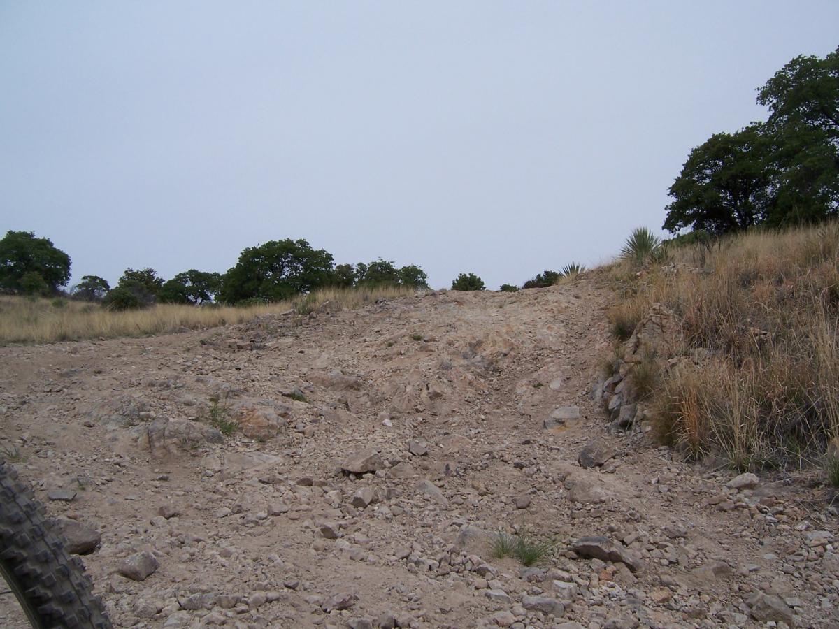 A rugged dirt path leading up a hillside, surrounded by sparse vegetation and low trees under a cloudy sky. The terrain is rocky and uneven, suggesting a natural outdoor environment. Brown Canyon mountain bike trail.