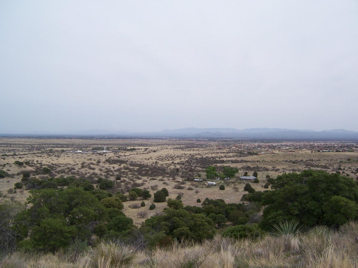 A panoramic view of a vast, open landscape featuring sparse vegetation and rolling hills under a cloudy sky. In the foreground, a few trees can be seen, while the background showcases distant mountains and a small cluster of buildings in a valley below. The scene conveys a sense of solitude and natural beauty. Brown Canyon mountain bike trail.