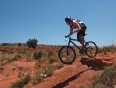 A person riding a mountain bike jumps off a rocky ledge in a desert landscape under a clear blue sky. The terrain features red soil and sparse vegetation. Palo Duro Canyon mountain bike trail.