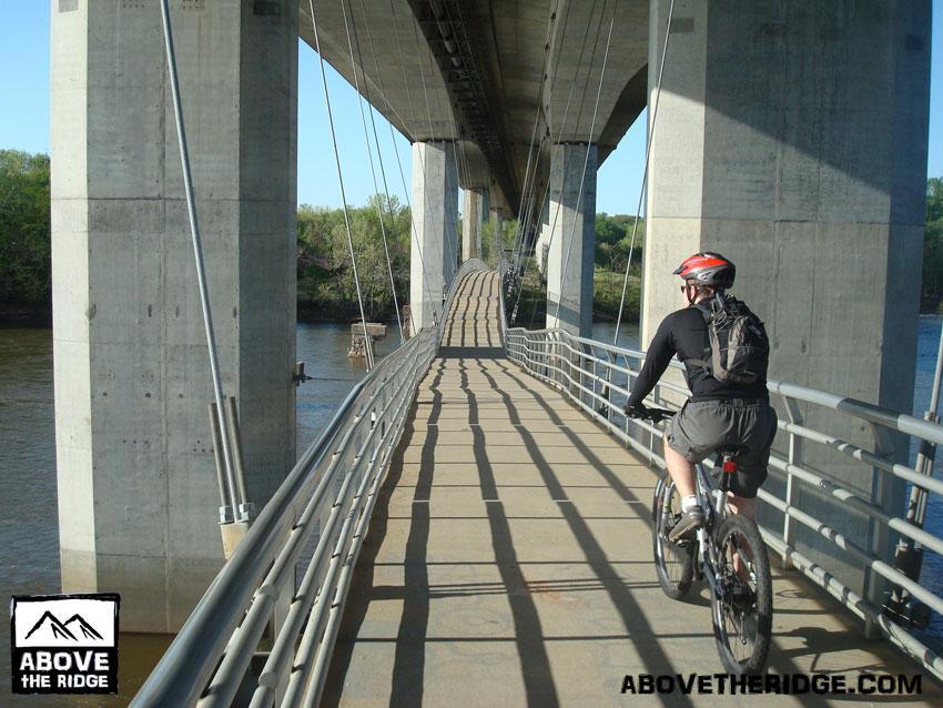 A cyclist riding on a narrow, elevated bike path underneath a large bridge, with concrete support columns and a river visible below. The scene is sunny, and the path features shadows cast by the bridge structure. Belle Isle mountain bike trail.