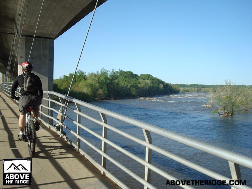 A cyclist riding a mountain bike along a bridge with a view of a river and green vegetation on the opposite bank. The scene is set on a sunny day, with clear blue skies and calm water flowing below. Belle Isle mountain bike trail.