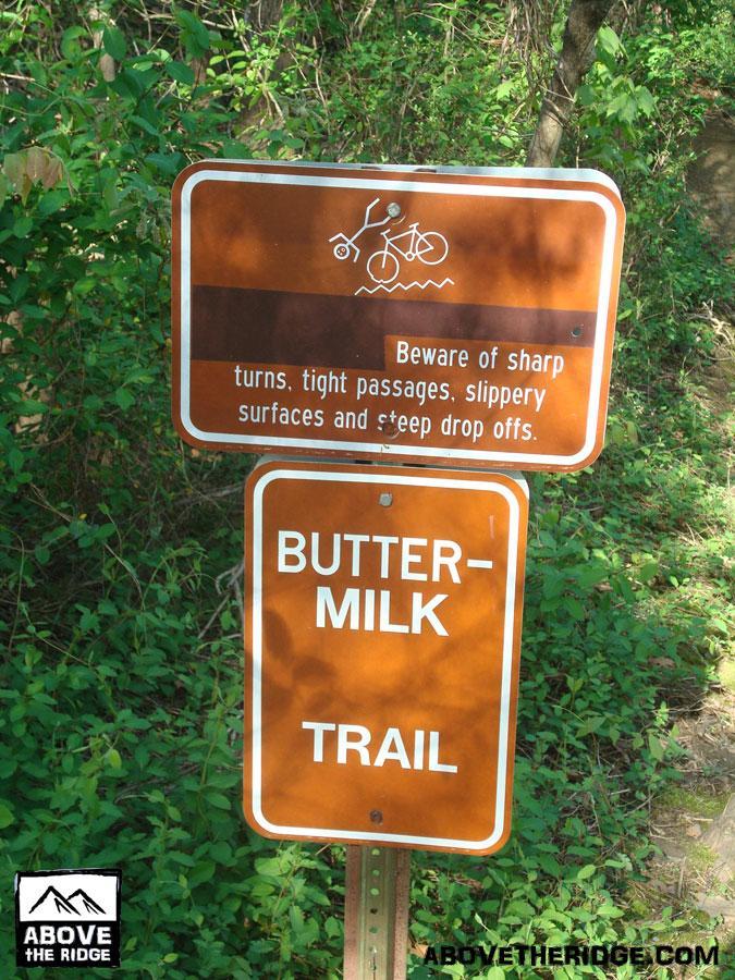 A trail sign indicating the "Buttermilk Trail," warning of sharp turns, tight passages, slippery surfaces, and steep drop-offs. The sign includes an illustration of a bicycle. The background features lush green foliage. Buttermilk mountain bike trail.