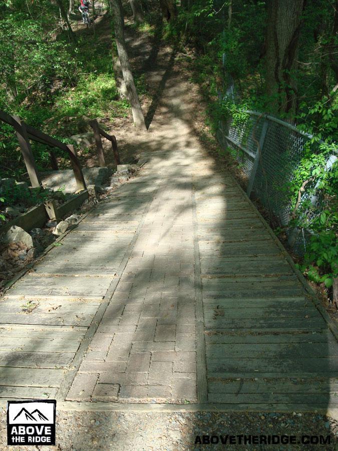 A downward path winding through a wooded area, featuring a wooden boardwalk with adjacent railings and greenery surrounding the trail. Sunlight filters through the trees, creating a dappled light effect on the ground. Buttermilk mountain bike trail.
