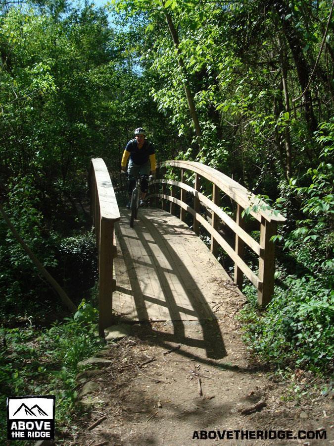 A person riding a mountain bike across a wooden bridge in a lush, green forest setting. Sunlight filters through the trees, casting shadows on the path. The environment features dense foliage and a dirt trail leading up to the bridge. Buttermilk mountain bike trail.