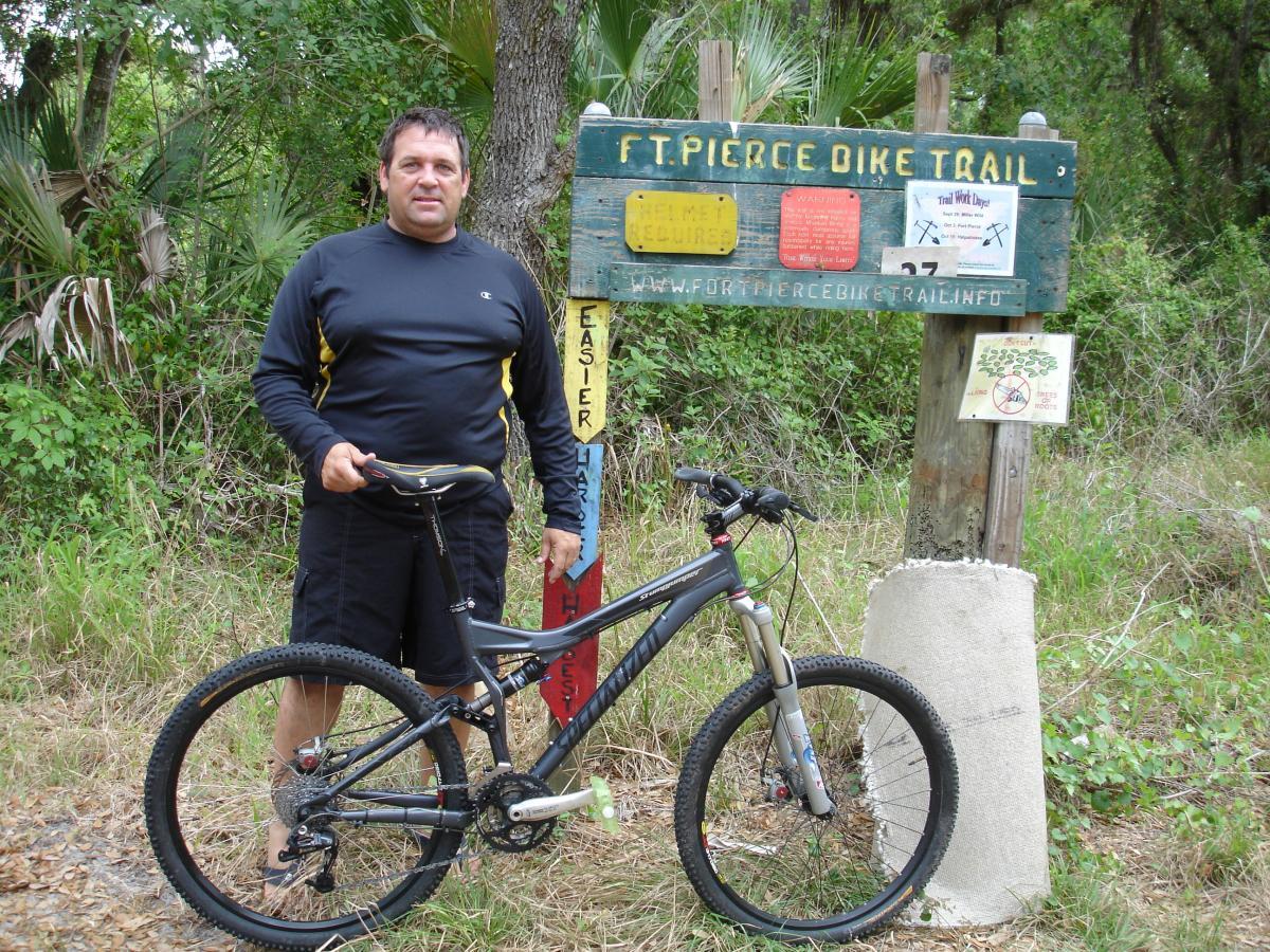 A man stands next to his mountain bike in front of the Fort Pierce Bike Trail sign. The sign includes various information about the trail, including difficulty levels and requirements. Surrounding him are trees and greenery typical of a biking trail environment. Fort Pierce Mountain Bike Trail mountain bike trail.