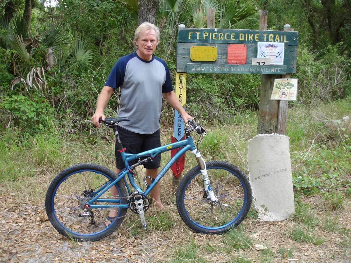 A person stands next to a blue mountain bike at the entrance of the Fort Pierce Bike Trail. The individual is wearing a gray and blue athletic shirt and shorts. Behind them is a wooden sign with trail information and difficulty levels. The background features lush greenery typical of a bike trail environment. Fort Pierce Mountain Bike Trail mountain bike trail.