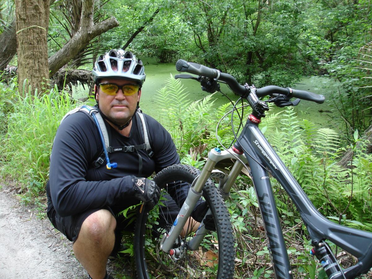 A man in a black long-sleeve shirt and sunglasses wearing a helmet kneels beside his mountain bike on a dirt trail surrounded by lush green vegetation and a body of water covered in algae. Fort Pierce Mountain Bike Trail mountain bike trail.