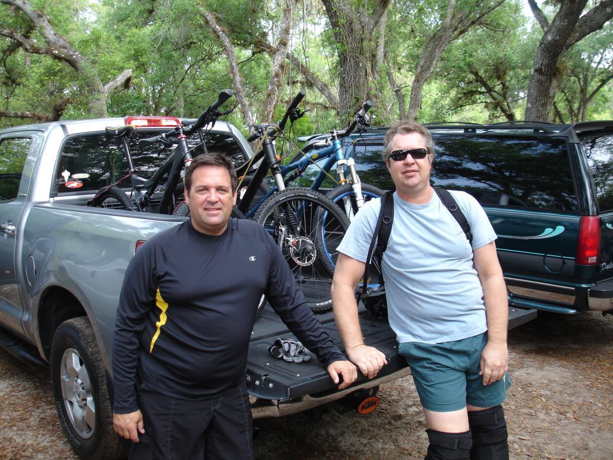 Two men stand by the back of a pickup truck, which is loaded with mountain bikes. The truck is parked in a wooded area, surrounded by trees. One man is wearing a black long-sleeve shirt and shorts, while the other is dressed in a light t-shirt, shorts, and sunglasses. Both appear to be preparing for a biking adventure. Fort Pierce Mountain Bike Trail mountain bike trail.