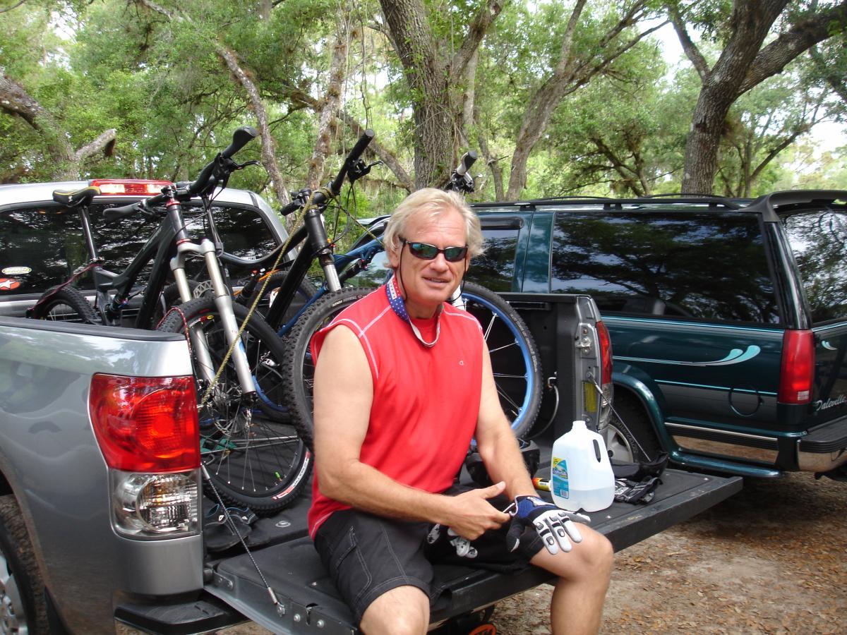 A man wearing sunglasses and a red tank top sits on the tailgate of a pickup truck, with several bicycles in the background. He is holding a mobile phone and has a water jug beside him. The setting is outdoors, surrounded by trees. Fort Pierce Mountain Bike Trail mountain bike trail.