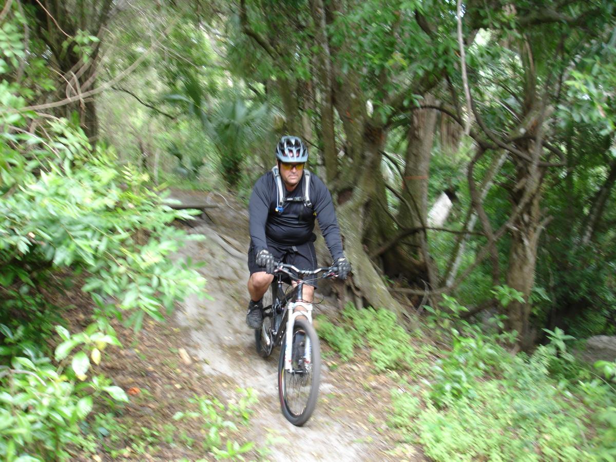A person riding a mountain bike on a narrow trail surrounded by lush greenery and trees. The cyclist is dressed in a black long-sleeve shirt, shorts, and wearing a helmet. The image captures the motion and excitement of biking in a natural environment. Fort Pierce Mountain Bike Trail mountain bike trail.