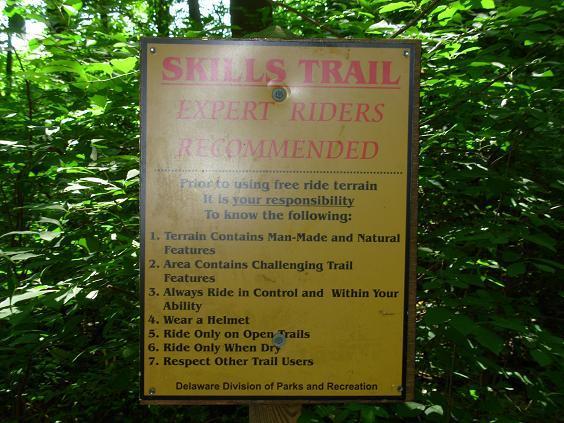 Signage for a skills trail in a wooded area, emphasizing that it is recommended for expert riders. The sign outlines important safety guidelines including wearing a helmet, riding in control, and respecting other trail users. The background features dense greenery typical of a forest setting. White Clay Creek mountain bike trail.