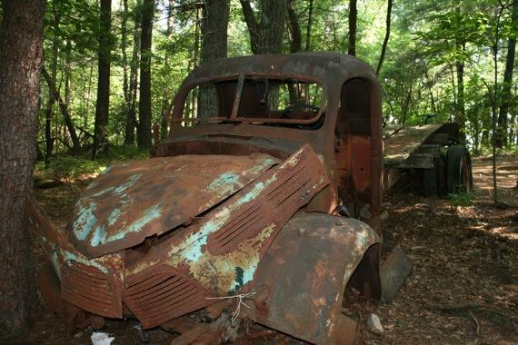 An old, rusty truck sits amidst trees in a forest setting, partially covered in foliage. The truck has a distressed appearance, with peeling paint, visible rust, and a damaged front end. The background features a lush forest environment, adding to the sense of neglect and abandonment. Bull / Jake Mountain mountain bike trail.