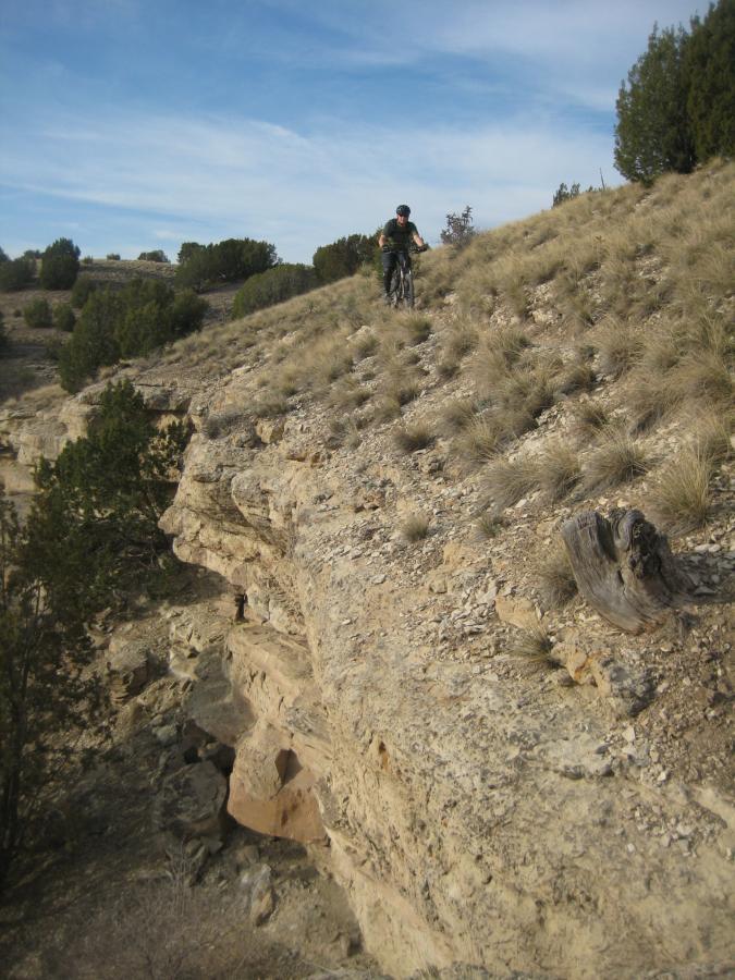 Mountain biker navigating a rocky terrain on a hillside, surrounded by sparse vegetation and a clear blue sky. The scene captures the adventurous spirit of outdoor cycling in a rugged landscape. South Shore Lake Pueblo mountain bike trail.