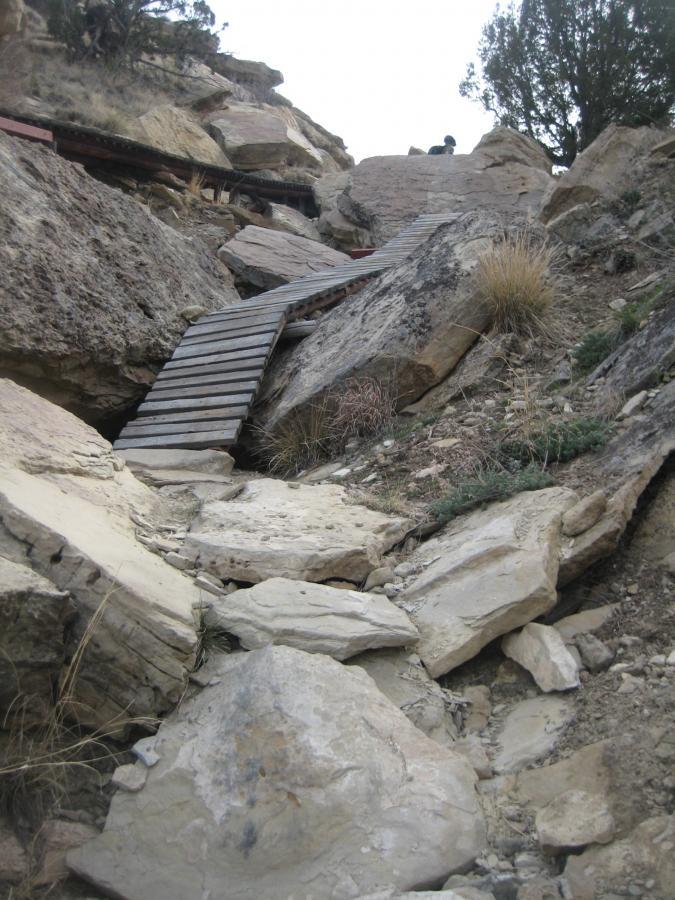 A rocky path leading up a steep incline, featuring a wooden walkway zigzagging between large boulders. Sparse vegetation is visible, and a person can be seen at the top of the rocks. The environment appears rugged and natural, with a mix of dirt, grass, and stone. South Shore Lake Pueblo mountain bike trail.