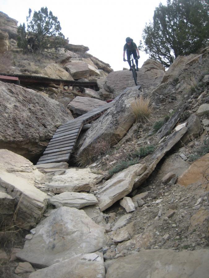 A mountain biker navigates a rocky trail featuring large boulders and a wooden bridge, set against a rugged landscape with sparse vegetation and overcast skies. South Shore Lake Pueblo mountain bike trail.