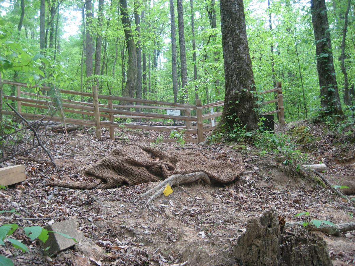 A wooded area featuring a path blocked by a wooden fence with a sign stating "CLOSED." In the foreground, a burlap sack is lying on the ground among leaves and dirt, with some exposed tree roots and small rocks visible. The surrounding environment is lush with green foliage typical of a forested area. Sope Creek mountain bike trail.
