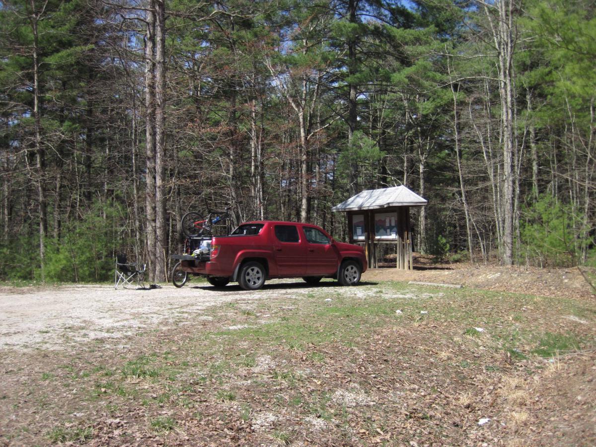 A red pickup truck parked on a gravel area near a nature trail. A folding chair is set up beside the truck, and a small covered information kiosk with a wooden structure is visible in the background, surrounded by trees. The scene is set in a wooded area with a mix of bare and green foliage, reflecting early spring. North Mountain/longdale Loop mountain bike trail.
