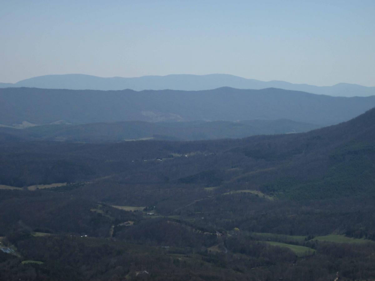 A panoramic view of rolling mountain ranges beneath a clear blue sky, showcasing layers of distant peaks and valleys with a mix of bare trees and patches of greenery. The landscape features varying shades of brown and green, indicating the transition of seasons. North Mountain/longdale Loop mountain bike trail.