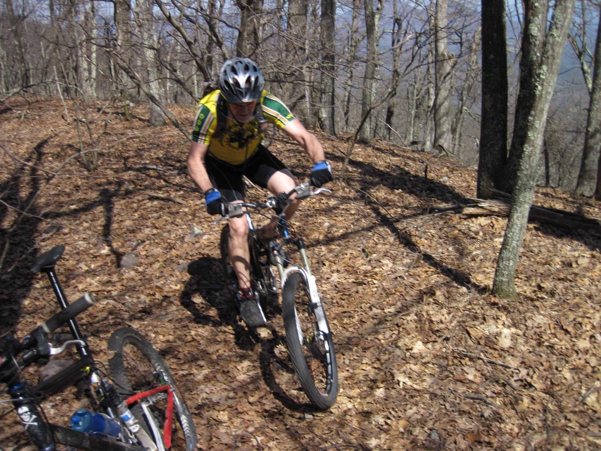 A mountain biker navigating a leaf-covered trail in a wooded area, wearing a yellow jersey and a helmet, focused on the ride. Another bicycle is partially visible in the foreground. North Mountain/longdale Loop mountain bike trail.