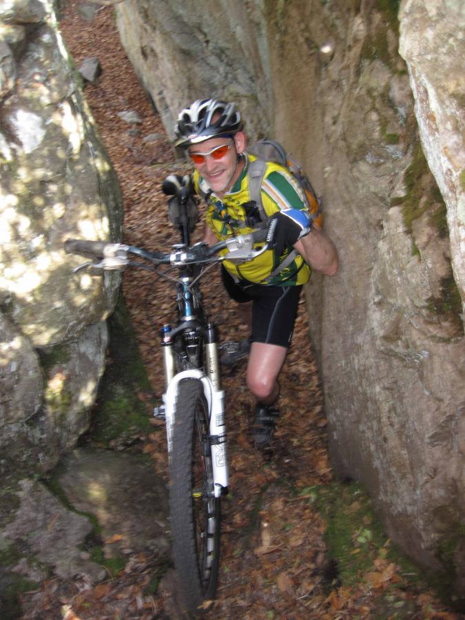 A mountain biker navigating a narrow gravel path between two large rock formations, smiling and wearing a helmet, sunglasses, and a colorful cycling jersey. Surrounding the path is a forested area with fallen leaves on the ground. North Mountain/longdale Loop mountain bike trail.