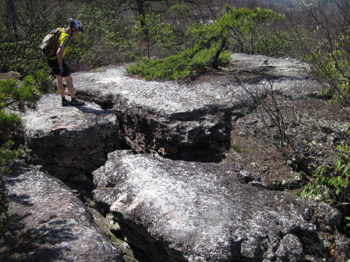 A person wearing a helmet and biking gear stands on a rocky ledge, looking down into a crevice surrounded by sparse vegetation. The scene is set in a natural outdoor environment, showcasing jagged rocks and a few small trees in the background. North Mountain/longdale Loop mountain bike trail.