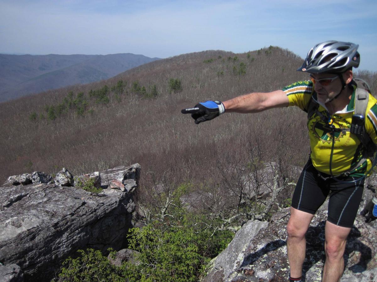 A cyclist in a yellow jersey and helmet points towards a distant landscape from a rocky vantage point on a mountain. The background features rolling hills covered in sparse trees, indicating a transition from winter to spring. The sky is clear, reflecting a bright, sunny day. North Mountain/longdale Loop mountain bike trail.
