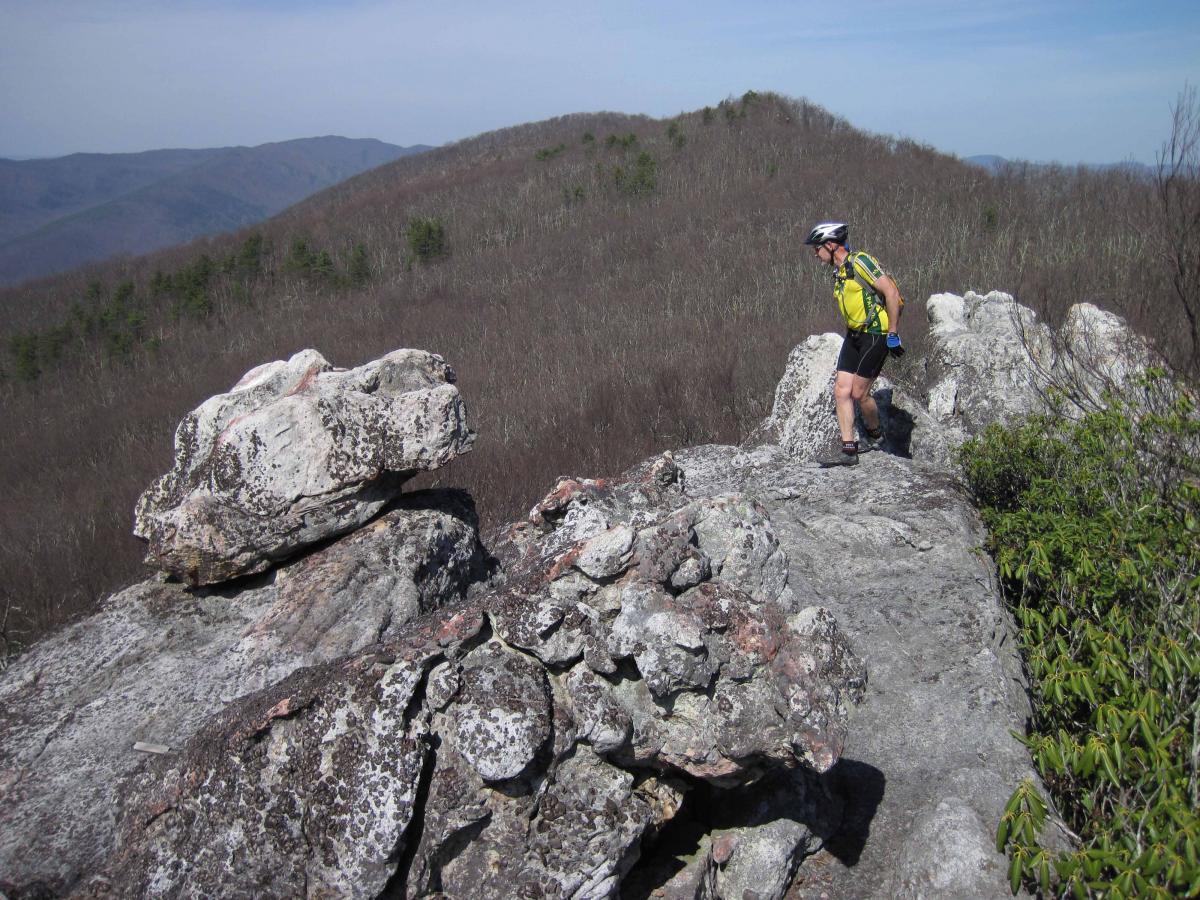 A person wearing a yellow shirt, black shorts, and a helmet is navigating a rocky outcrop on a mountain trail. The landscape features rolling hills in the background, with sparse trees and a clear blue sky. North Mountain/longdale Loop mountain bike trail.