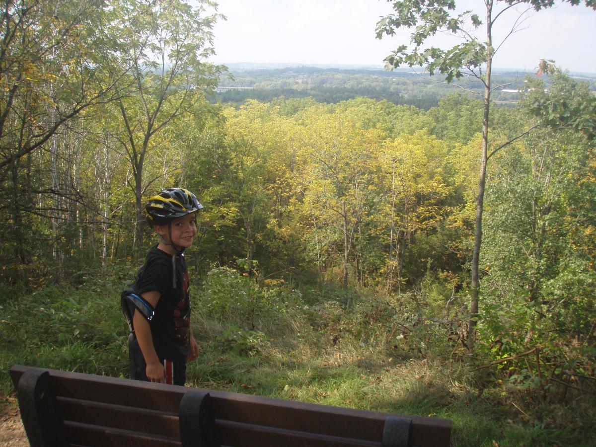 A young boy wearing a helmet stands on a hillside, smiling and looking back at the camera. He is in a forested area with vibrant autumn foliage in shades of yellow and green. In the foreground, there is a wooden bench, and the view in the background shows a sprawling landscape of trees and gentle hills under a blue sky. John Muir Trails mountain bike trail.