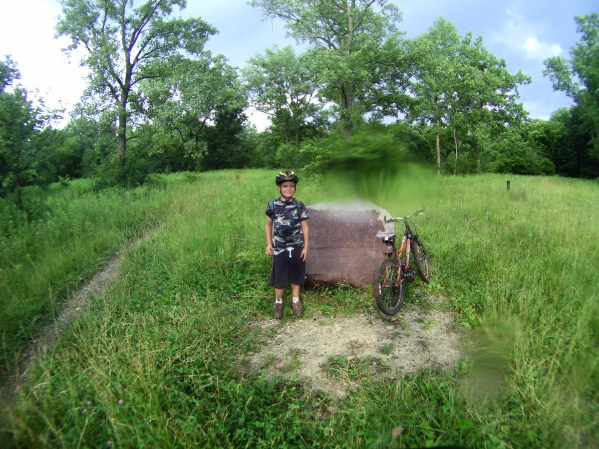 A young boy wearing a helmet and a camouflage shirt stands beside a bicycle in a lush green field, with trees in the background. He is smiling and poses in front of a large rock feature partially obscured by grass. Palos Forest Preserve mountain bike trail.