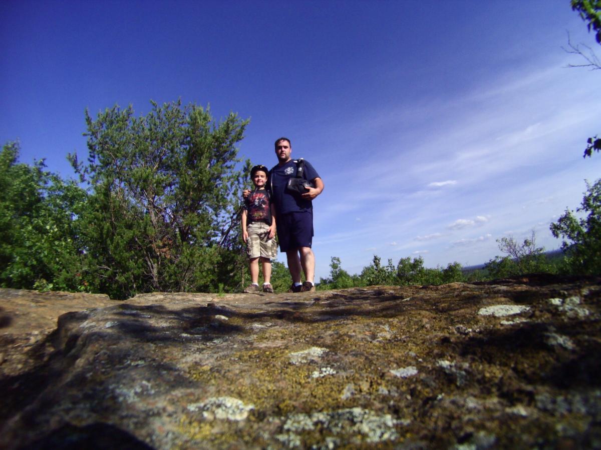 A man and a child standing on a rocky ledge with greenery in the background under a clear blue sky. The man is wearing a dark shirt and shorts, while the child is in a colorful t-shirt and shorts, wearing a hat. Levis Mounds mountain bike trail.