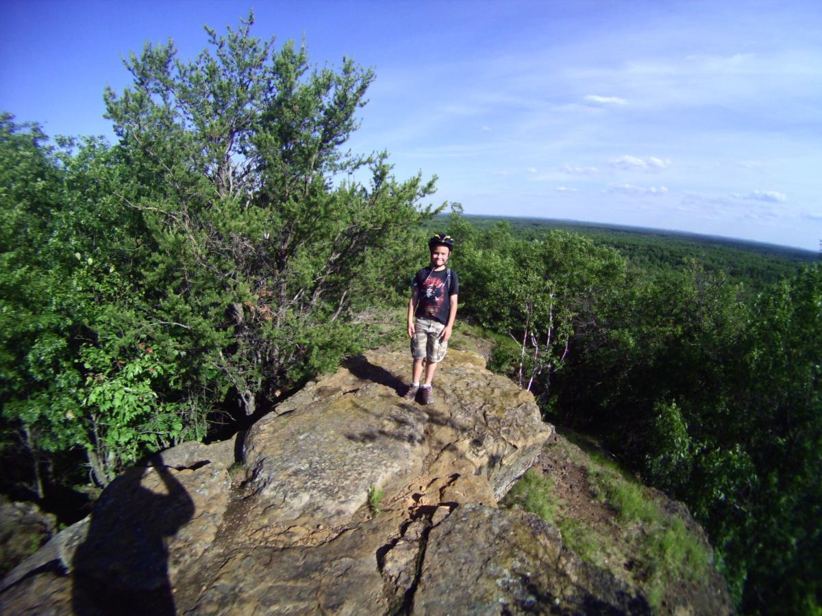 A young person standing on a rocky ledge surrounded by green trees, with a clear blue sky in the background. The landscape stretches out into the distance, showcasing a vast expanse of forest and horizon. The individual is smiling and wearing casual clothes, including a helmet. Levis Mounds mountain bike trail.