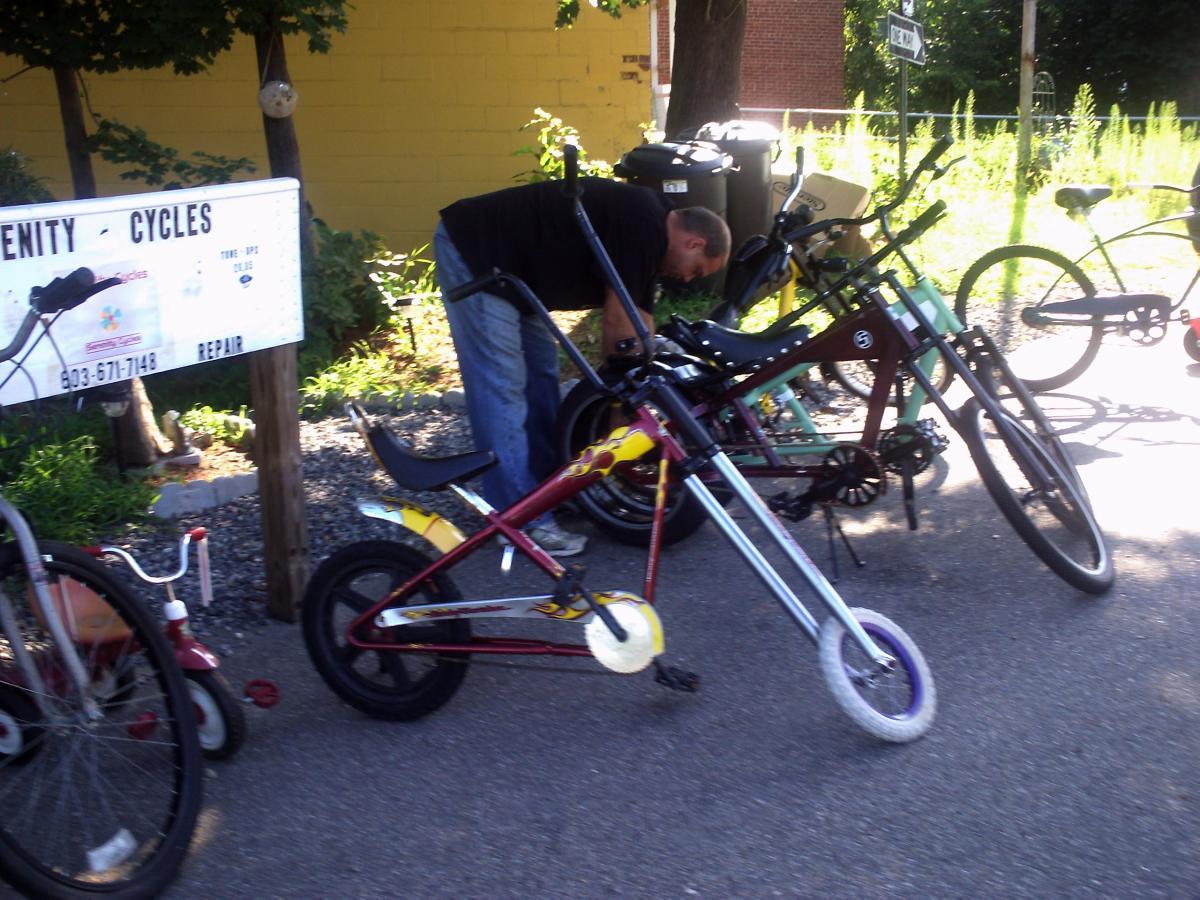 A person is repairing a bicycle in front of a sign that reads "Community Cycles," with various bicycles parked nearby. The scene is set outdoors, with greenery and a yellow building in the background.