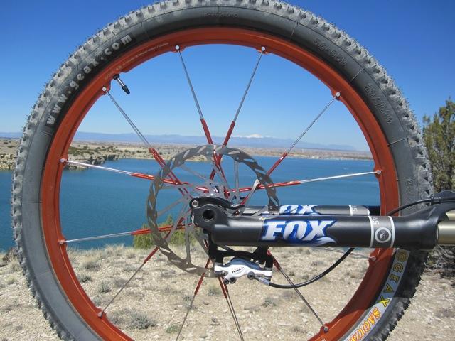 Close-up view of a mountain bike wheel featuring an orange rim, black tire, and visible disc brake. In the background, a serene blue lake and distant mountains under a clear sky can be seen. South Shore Lake Pueblo mountain bike trail.