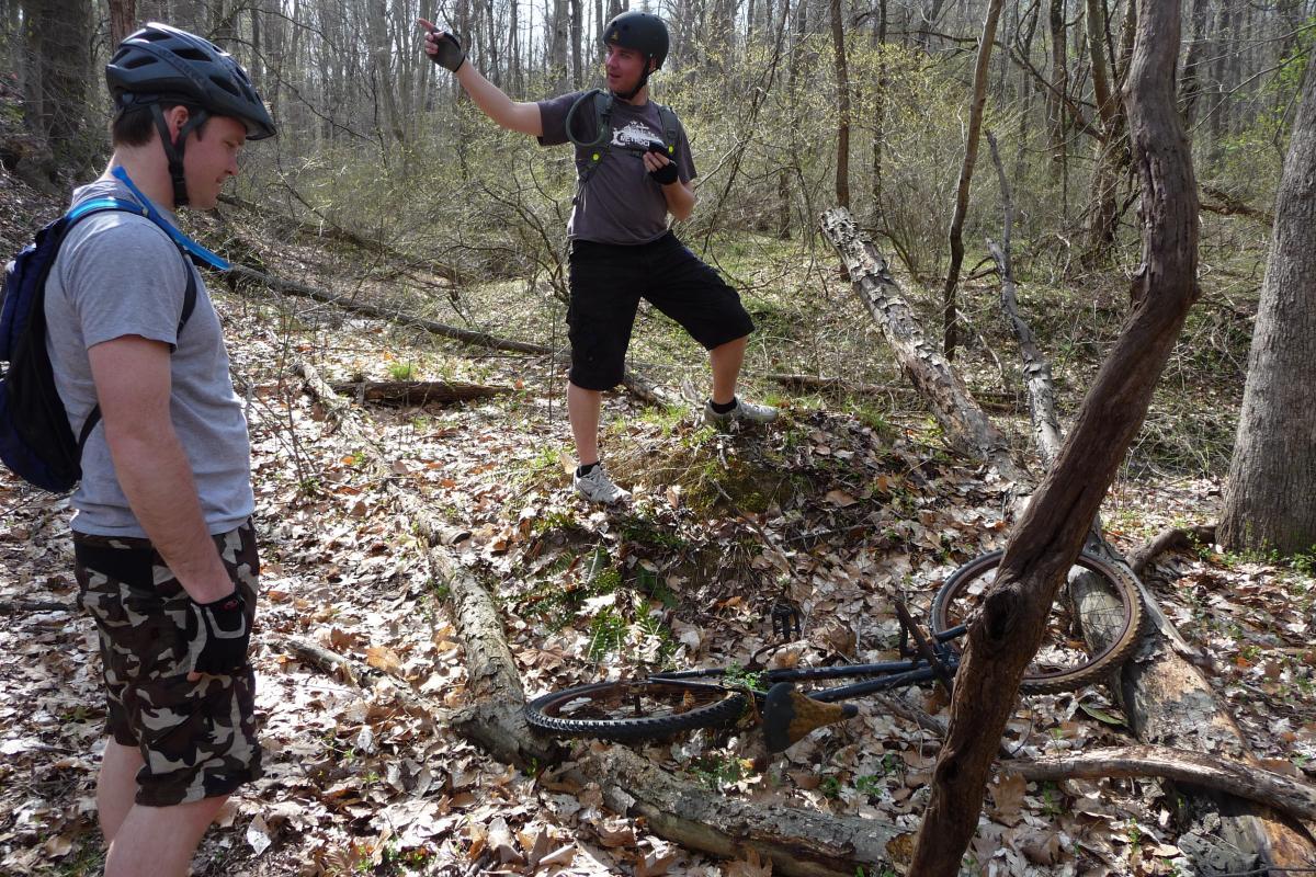 Two men in helmets are in a forested area. One man stands on a small mound with a bike resting on the ground among fallen leaves and logs, while he appears to be looking at a camera or phone. The second man stands nearby, looking down at the ground, wearing a t-shirt and camouflage shorts. The scene captures a moment during a mountain biking or outdoor adventure. Rosaryville State Park mountain bike trail.