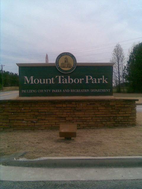 Sign at the entrance of Mount Tabor Park, featuring the name of the park and the designation of the Paulding County Parks and Recreation Department, set against a natural landscape with trees in the background. Mt Tabor Park mountain bike trail.