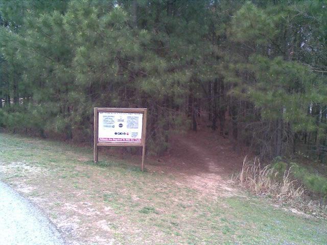 A pathway leading into a wooded area, marked by a wooden sign displaying information and rules. The foreground features grassy terrain and a gravel path, while dense trees provide a natural backdrop. Mt Tabor Park mountain bike trail.