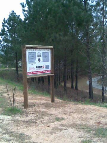 A wooden sign at the entrance to a natural area, surrounded by tall pine trees. The sign displays information and guidelines for visitors, including safety notices. A gravel path leads towards the sign, with a glimpse of a road in the background. Mt Tabor Park mountain bike trail.