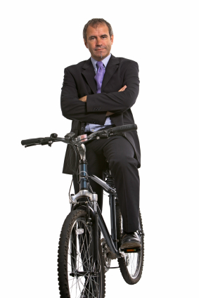 A businessman in a suit and tie confidently poses while sitting on a mountain bike, with his arms crossed and a slight smile, against a white background.