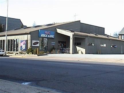 A bicycle shop with a large sign reading "Bike Line" on the front. The store features large windows showcasing various bicycles inside. The building is gray and has a modern architectural design, with a wide entrance and several bikes displayed outside. The scene captures the shop from across the street.