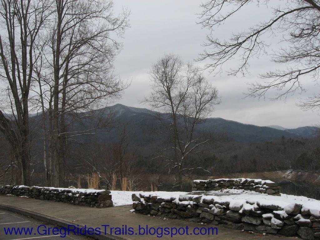 A scenic view of a mountainous landscape with bare trees in the foreground, a stone wall along a pathway, and a cloudy sky. Snow is visible on the ground, indicating winter conditions. The mountains in the background are partially shrouded in mist, creating a serene and tranquil atmosphere. Fontana Village mountain bike trail.