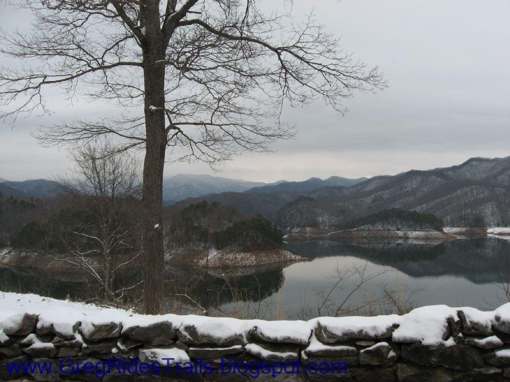A serene winter landscape featuring a leafless tree in the foreground, snowy mountains in the background, and a calm lake reflecting the surroundings. A stone wall is visible at the bottom of the image. The sky is overcast, adding a muted atmosphere to the scene. Fontana Village mountain bike trail.