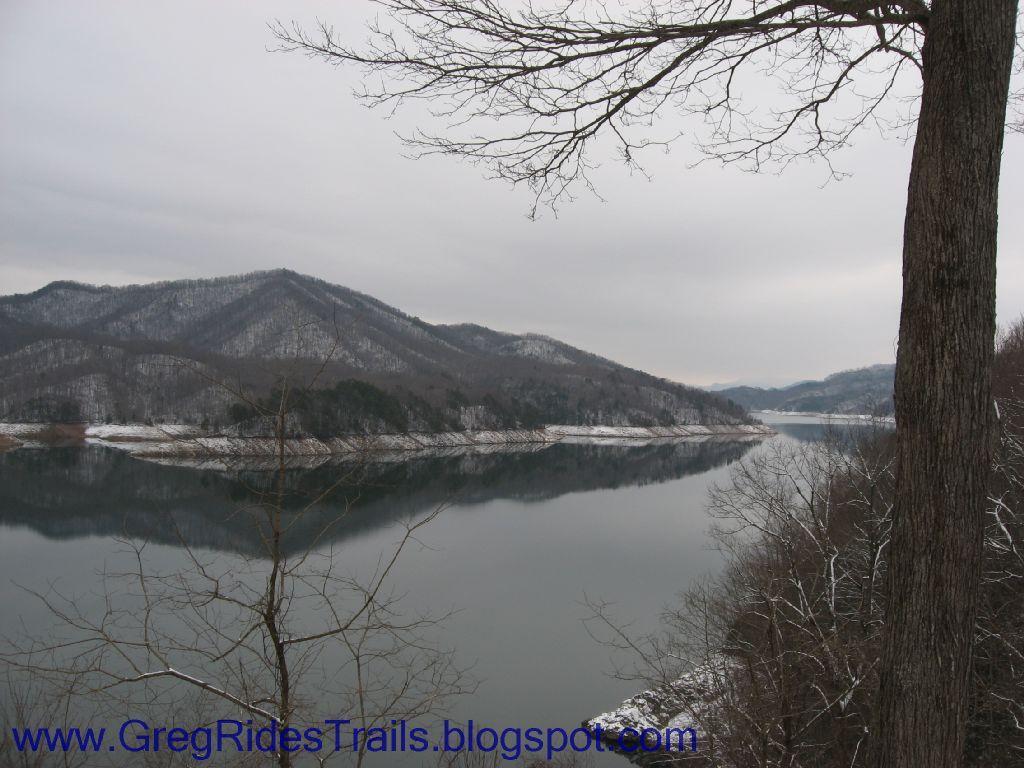 A scenic view of a calm lake surrounded by snow-capped mountains under a cloudy sky. The water reflects the landscape, creating a serene atmosphere, with leafless trees framing the image in the foreground. Fontana Village mountain bike trail.