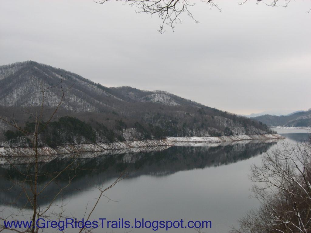 A serene landscape featuring a calm lake surrounded by snow-dusted hills under a cloudy sky. The reflection of the hills on the water creates a tranquil scene, with bare trees in the foreground. Fontana Village mountain bike trail.