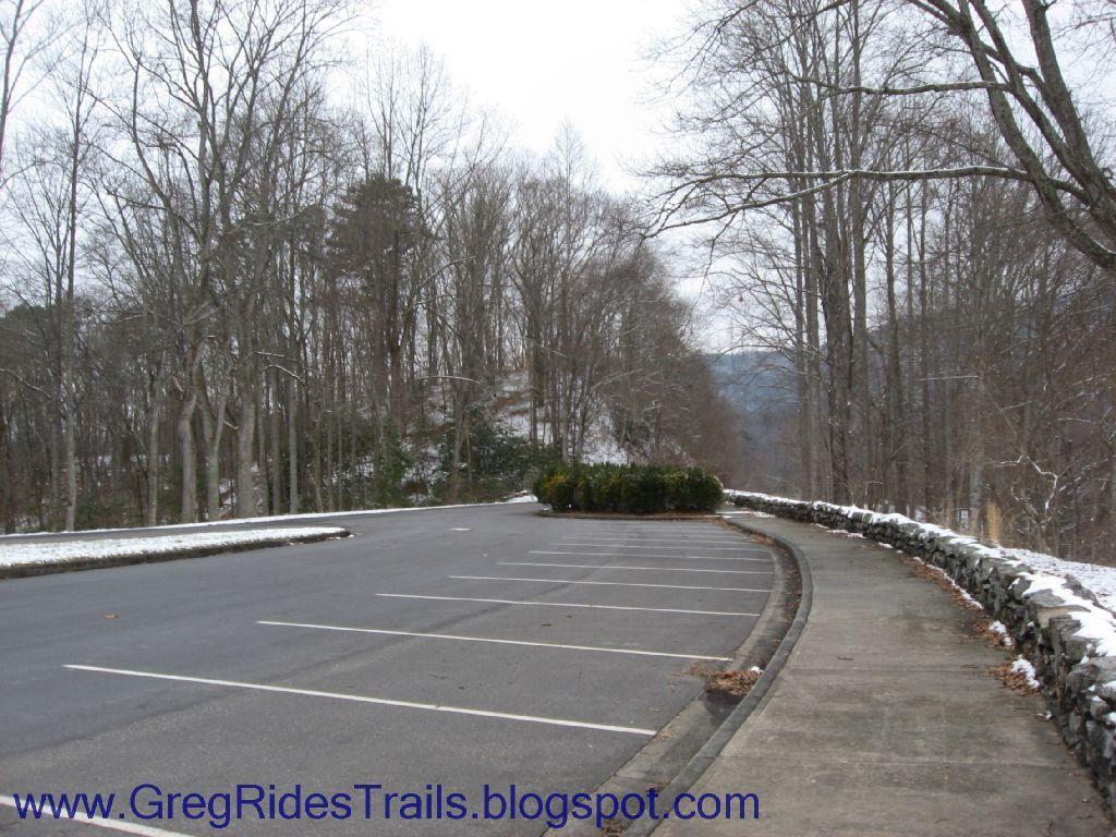 A quiet winter scene depicting an empty parking lot bordered by bare trees and a stone wall. Light snow covers the ground, creating a serene atmosphere. In the distance, hills are partially visible through the trees, suggesting a rural or wooded area. Fontana Village mountain bike trail.