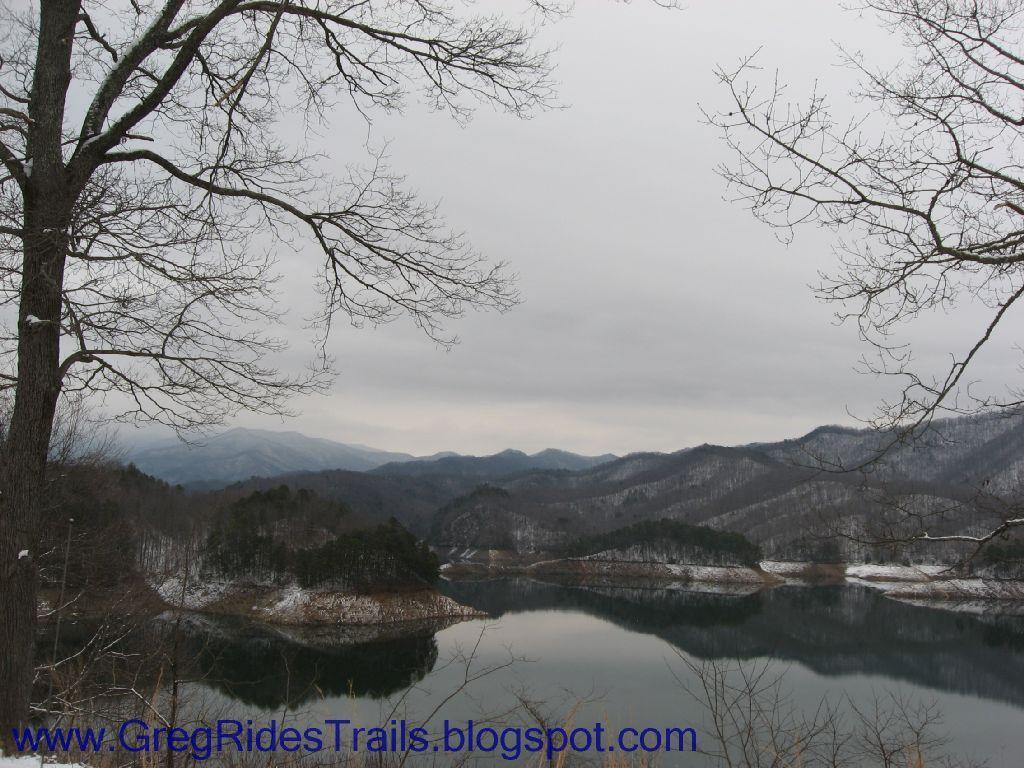 A tranquil winter landscape featuring a calm lake surrounded by rolling hills and mountains. The scene is framed by bare trees in the foreground, under a cloudy sky. Snow-capped peaks are visible in the distance, reflecting subtly in the still water. Fontana Village mountain bike trail.