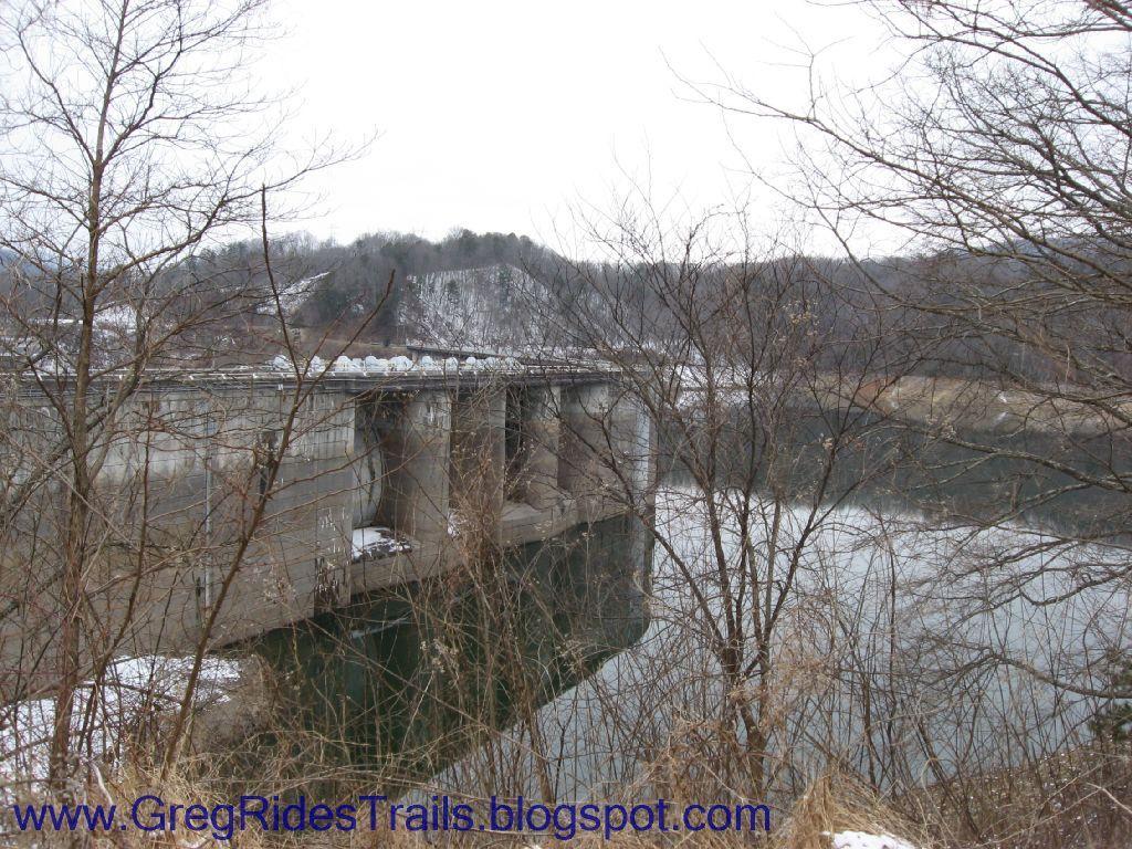 A concrete bridge spans a calm river, surrounded by bare trees and rolling hills covered in patches of snow. The sky is overcast, contributing to the chilly atmosphere of the scene. Fontana Village mountain bike trail.