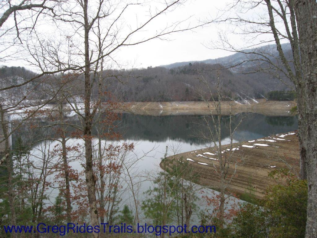A tranquil winter landscape featuring a calm lake surrounded by bare trees and distant mountains. The water reflects the overcast sky, and patches of snow can be seen on the ground, suggesting a serene, chilly day in nature. Fontana Village mountain bike trail.
